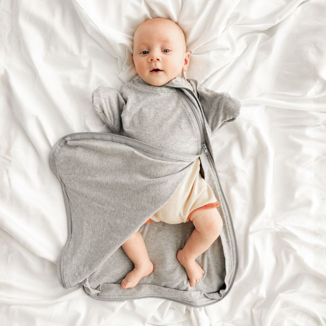 A baby lying on white bedding, wearing a gray sleep sack that has clever zippers for best nappy changing.