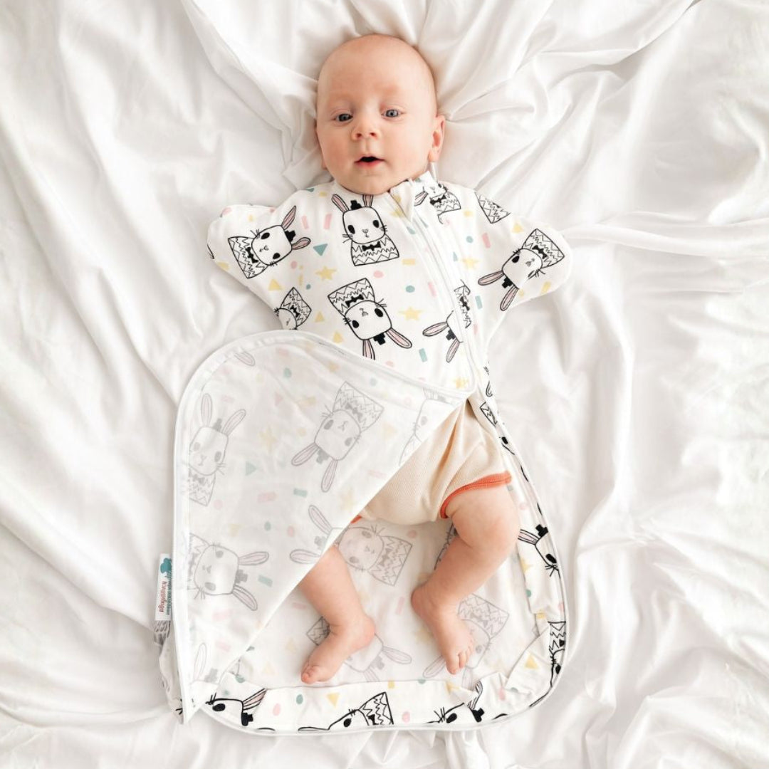 A baby lying on white bedding, wearing a white sleep sack with a black and gray bunny pattern.