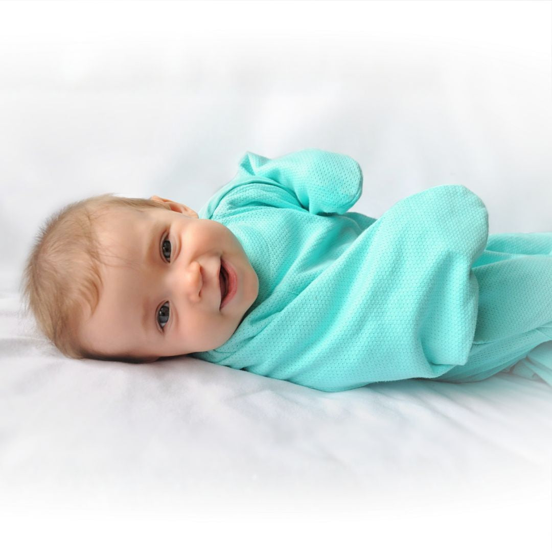 A happy baby lying on their side wearing a light aqua textured transitioning sleep swaddle.